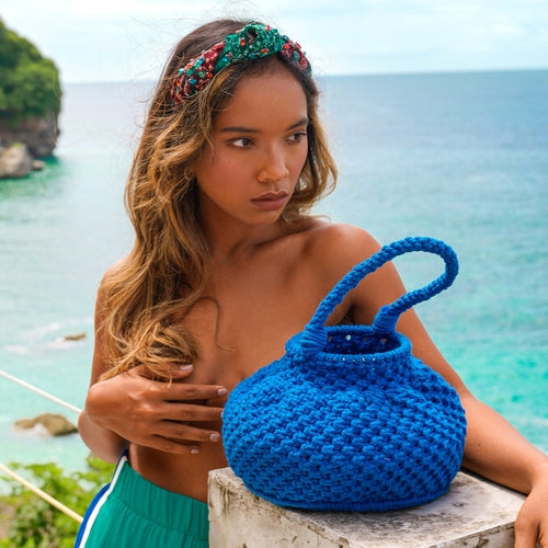 Azure blue macrame bucket bag displayed by a model against a scenic ocean backdrop