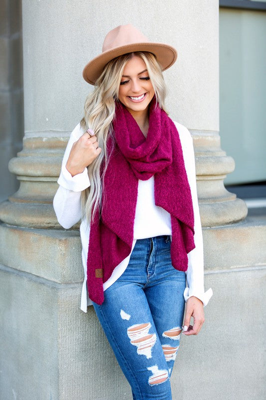 Woman wearing magenta cc cozy draped oversized scarves for women with white sweater ripped jeans tan hat leaning on stone column