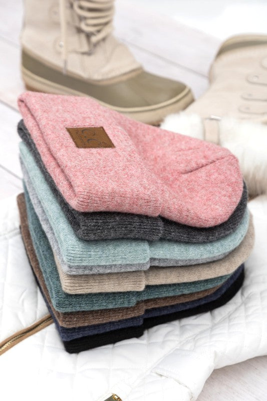 Stack of assorted knit CC heathered boyfriend beanie styles with leather patch displayed with winter boots