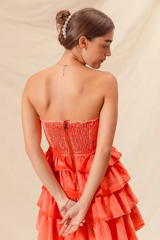 Back view of a woman wearing a strapless ruffle layered dress in bright coral, featuring a smocked bodice