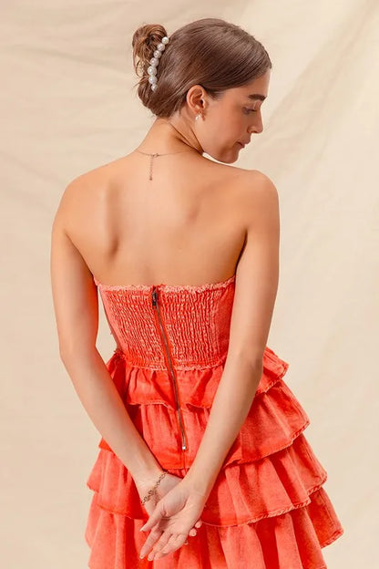 Back view of a woman wearing a strapless ruffle layered dress in bright coral, featuring a smocked bodice